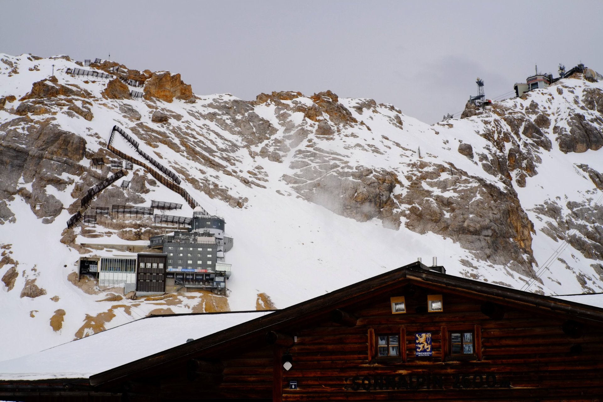 A photo of a dark wet wooden building, a high vertical cliff behind it, and a bunch of boxy buildings built into the cliff.