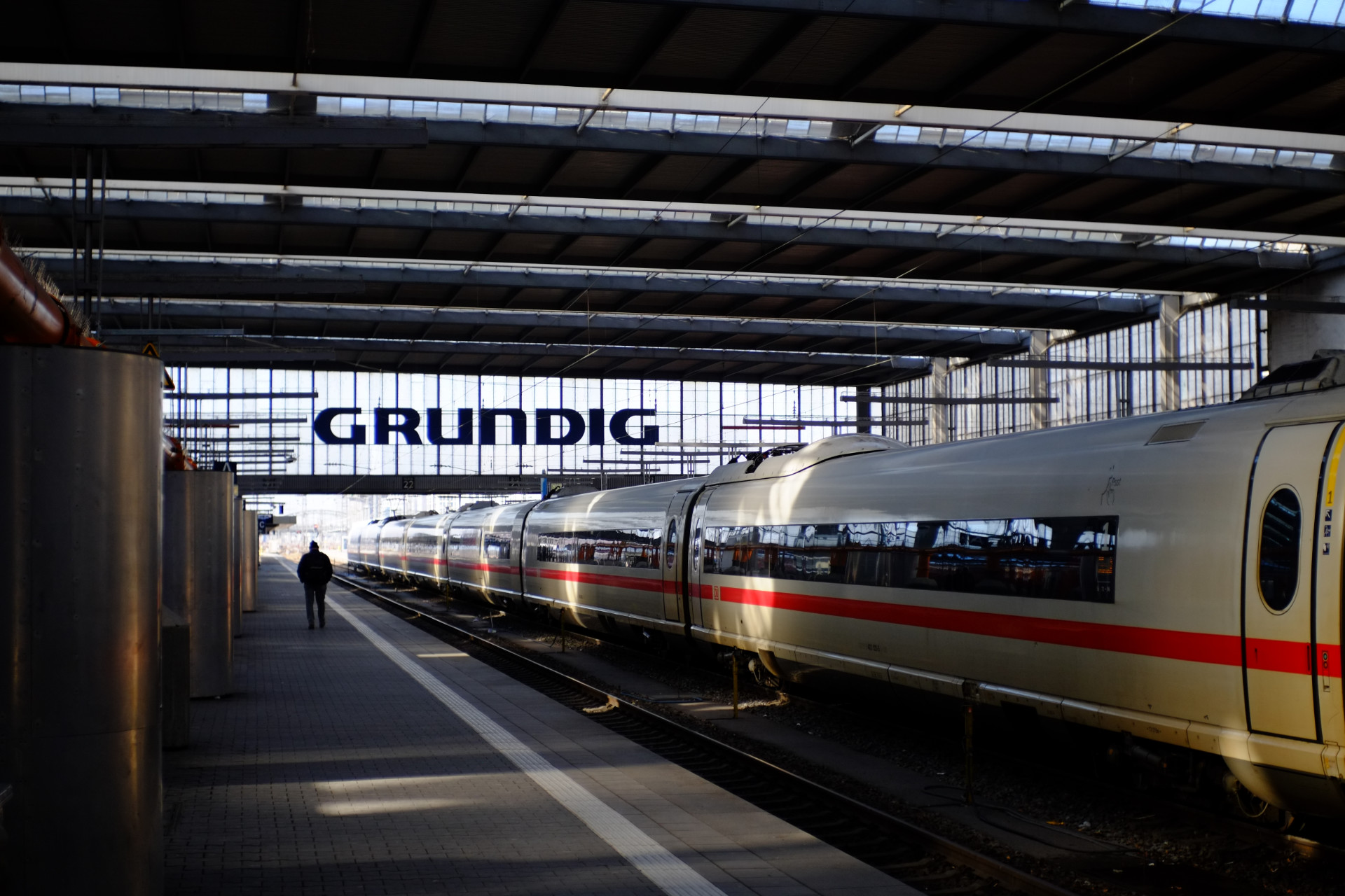 A photo of one of the München central train station's platforms featuring a white high-speed train with a thin red horizontal line drawn on every train car. In the background there are big letters on a glass wall saying: GRUNDIG.