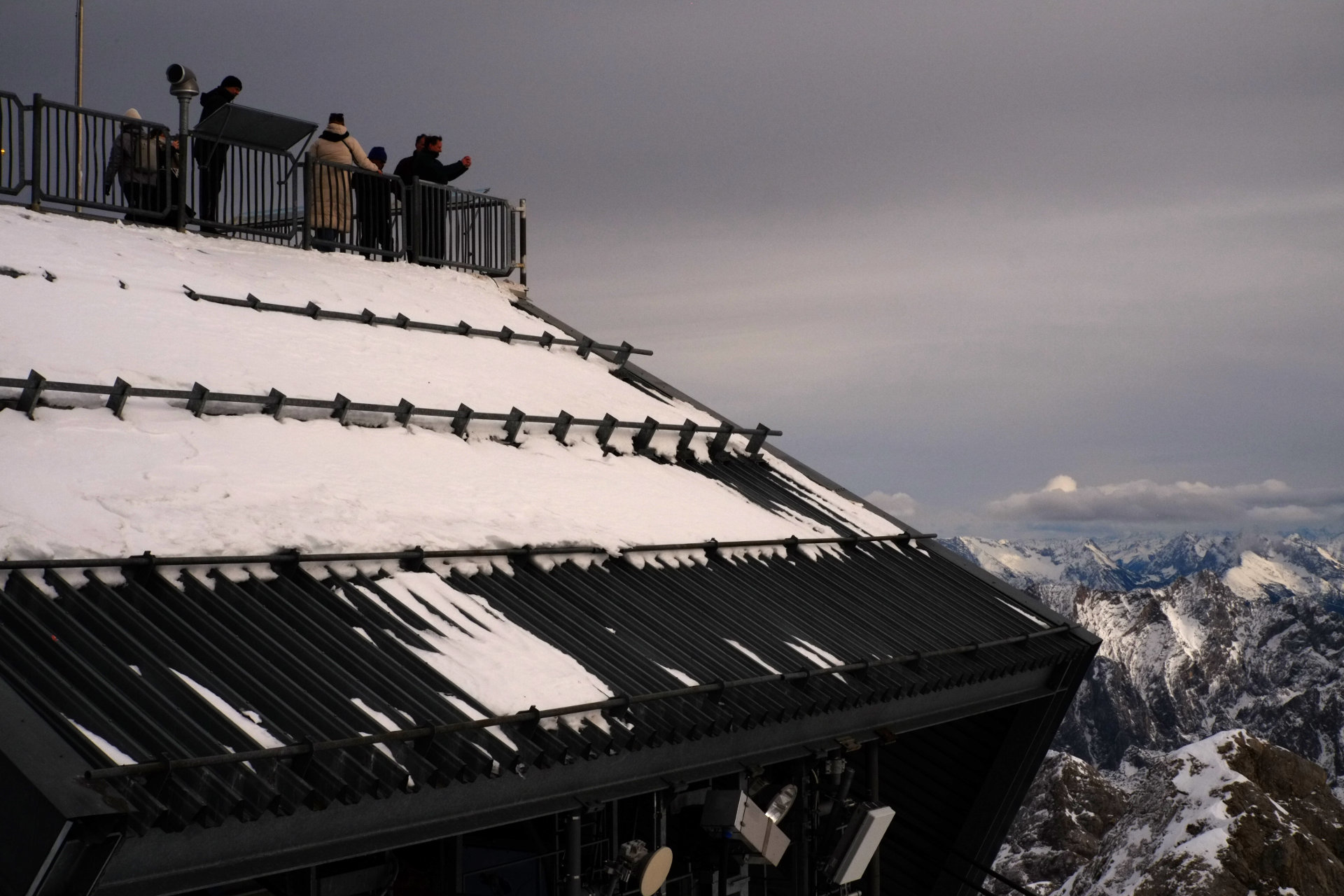 A photo of people staying at the observation deck with a roof slope under them and snowy mountains in the background.