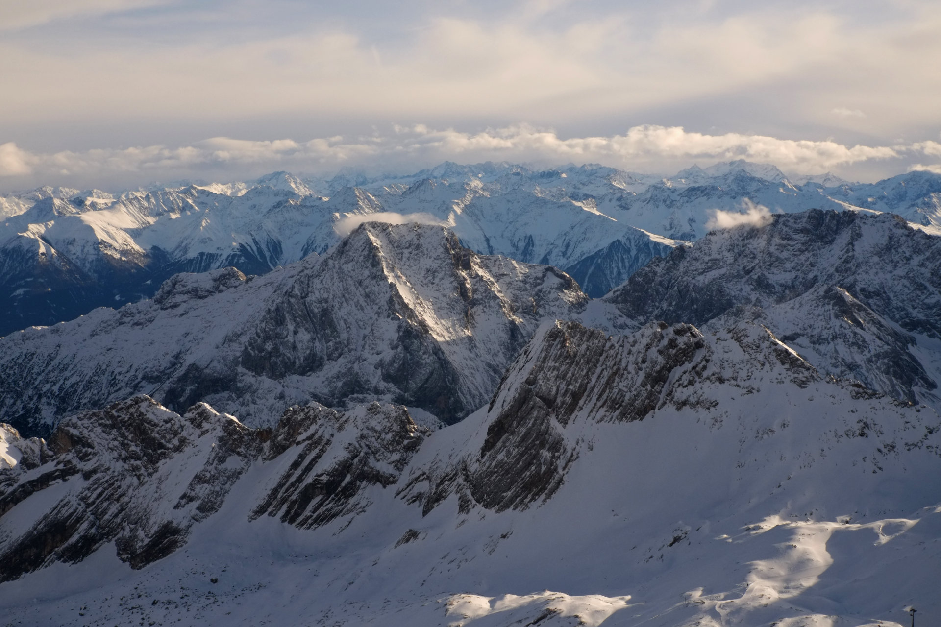 A photo of snowy mountain peaks illuminated by the evening sun and mountains behind them covered in blue shadows.
