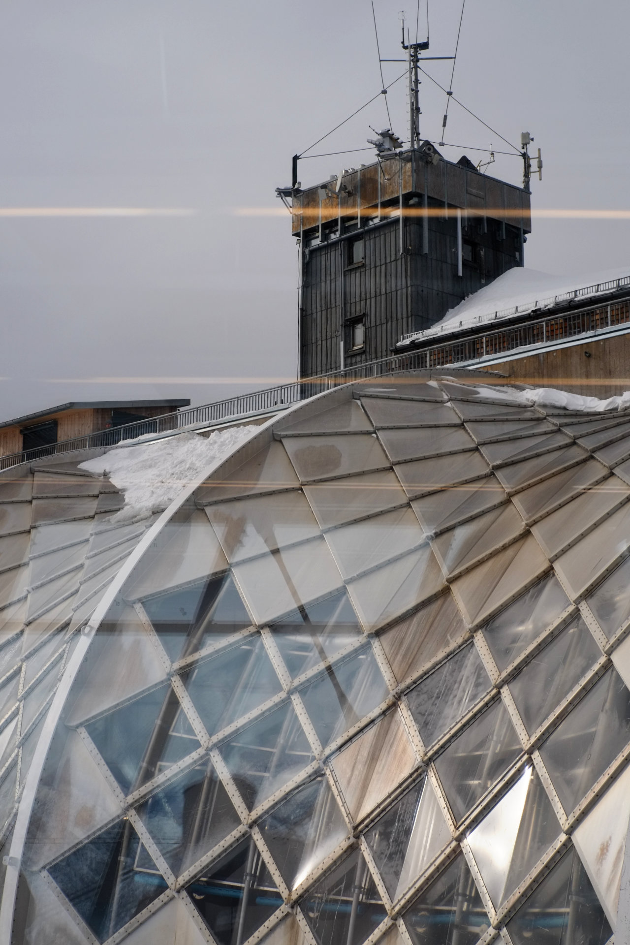 A photo of a cylindrical glass roof and a dark square tower on a platform behind and above the roof.