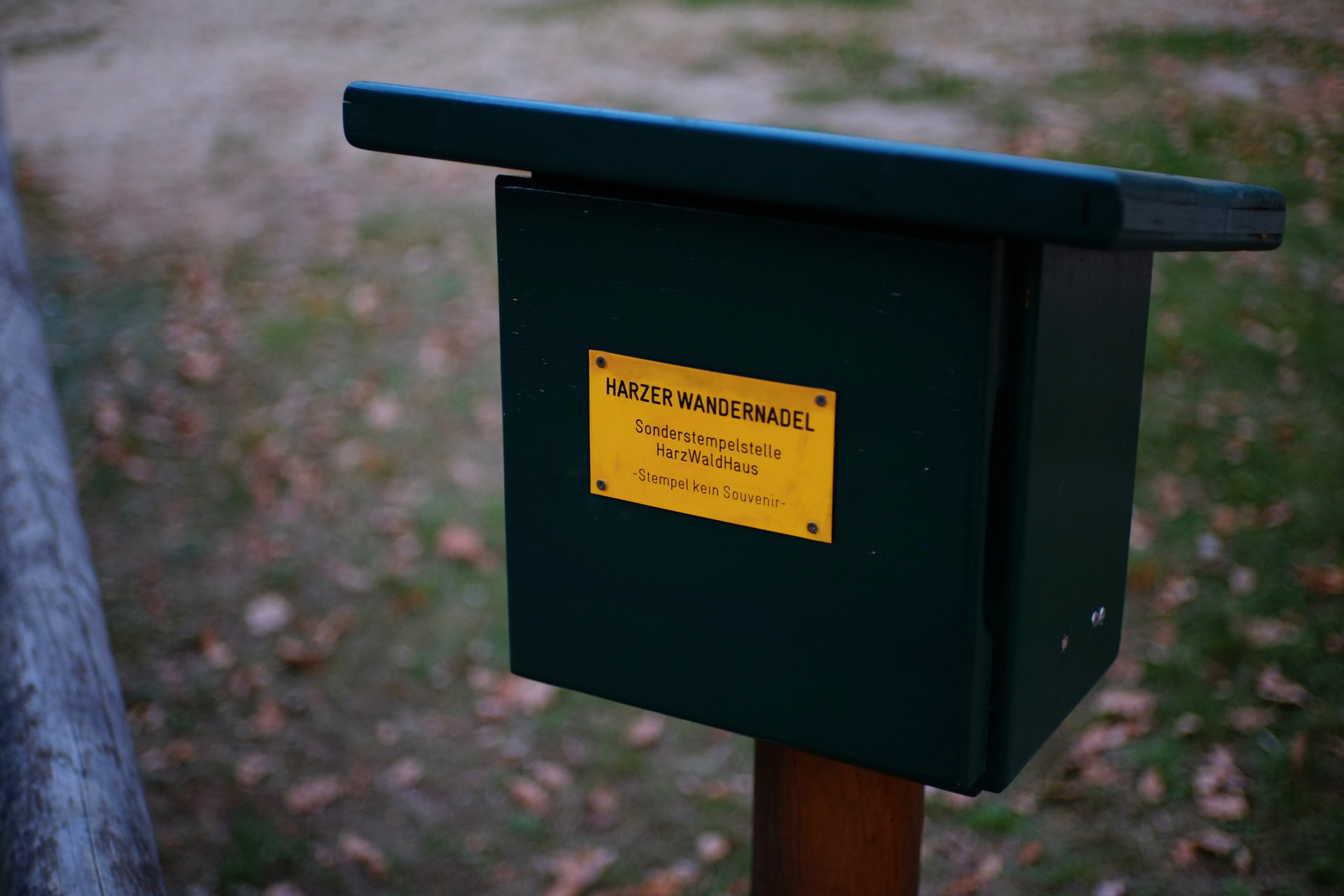 A photo of a green wooden box on a brown post with a yellow plaque saying "Harzer Wandernadel", "Sonderstempelstelle HarzWaldHaus", "Stempel kein Souvenir"