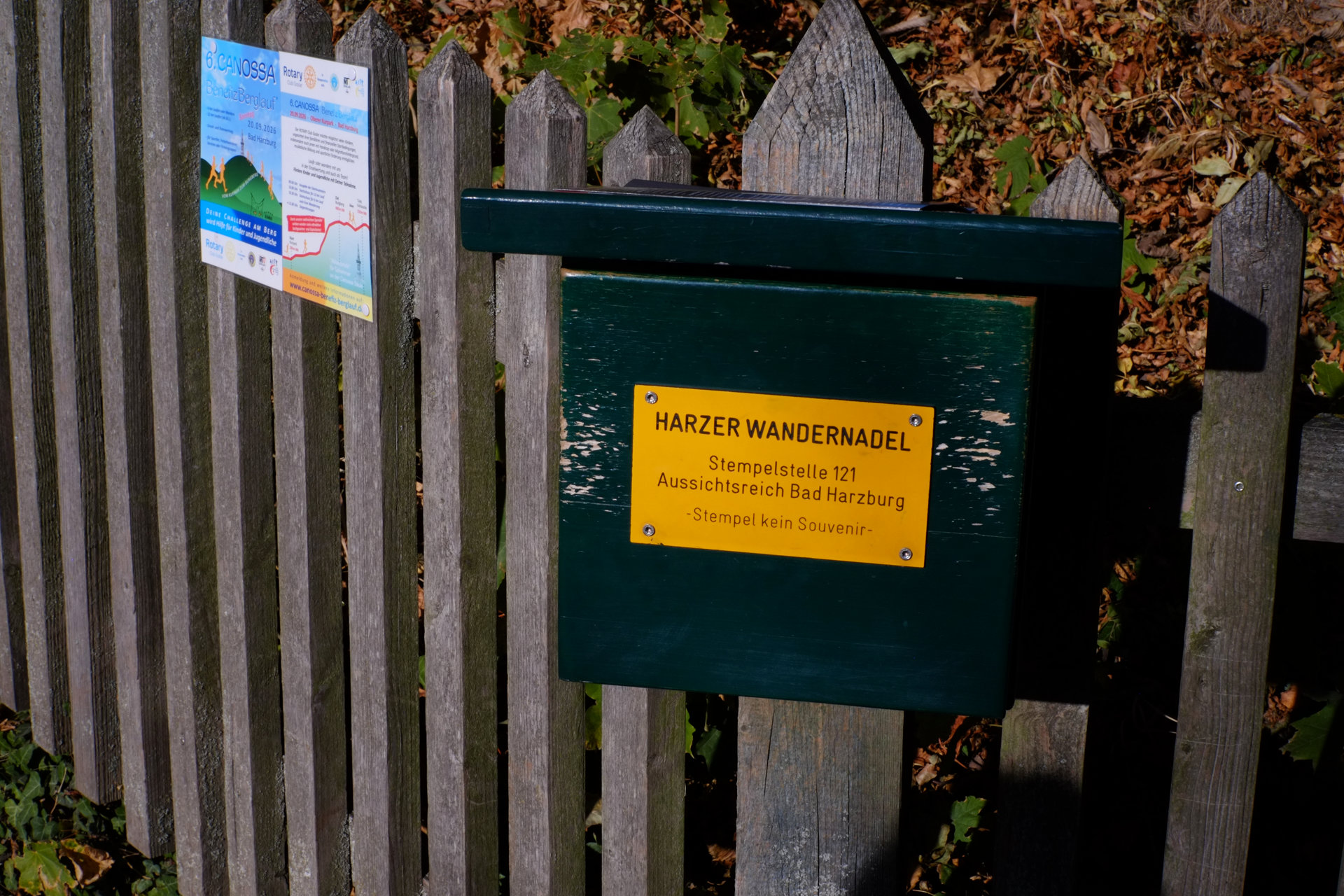 A photo of a green wooden box that is bolted to a dooden fence with a yellow plaque saying "Harzer Wandernadel", "Stempelstelle 121 Aussichtsreich Bad Harzburg", "Stempel kein Souvenir"