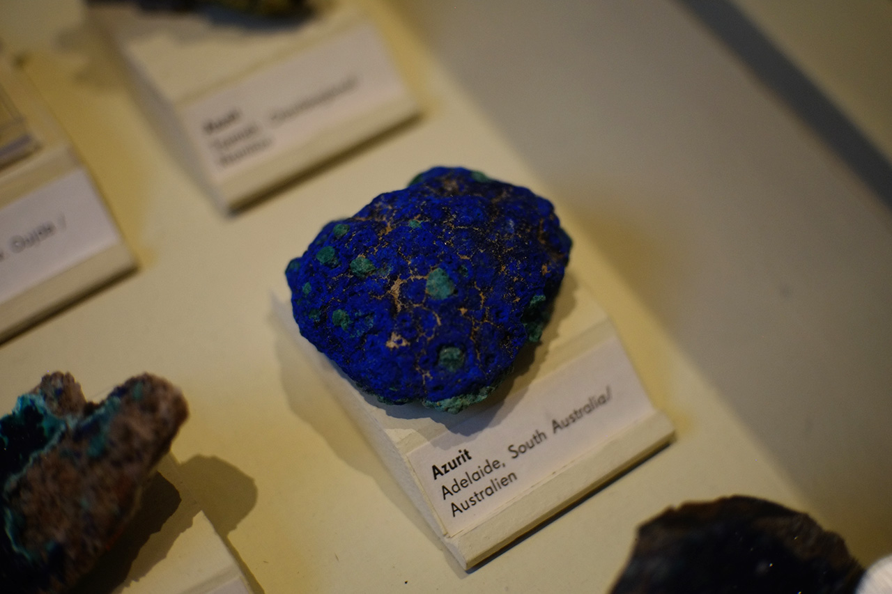 A photo of a deep blue mineral stone with green spots and beige veins exhibited under the glass on a table among other minerals. The label under it says: Azurit. Adelaide, South Australia /Australien