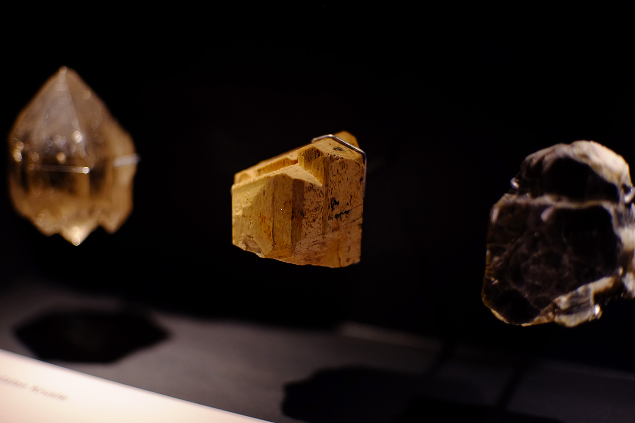 A photo of three small minerals exhibited under the light on a shelf with black back sides.