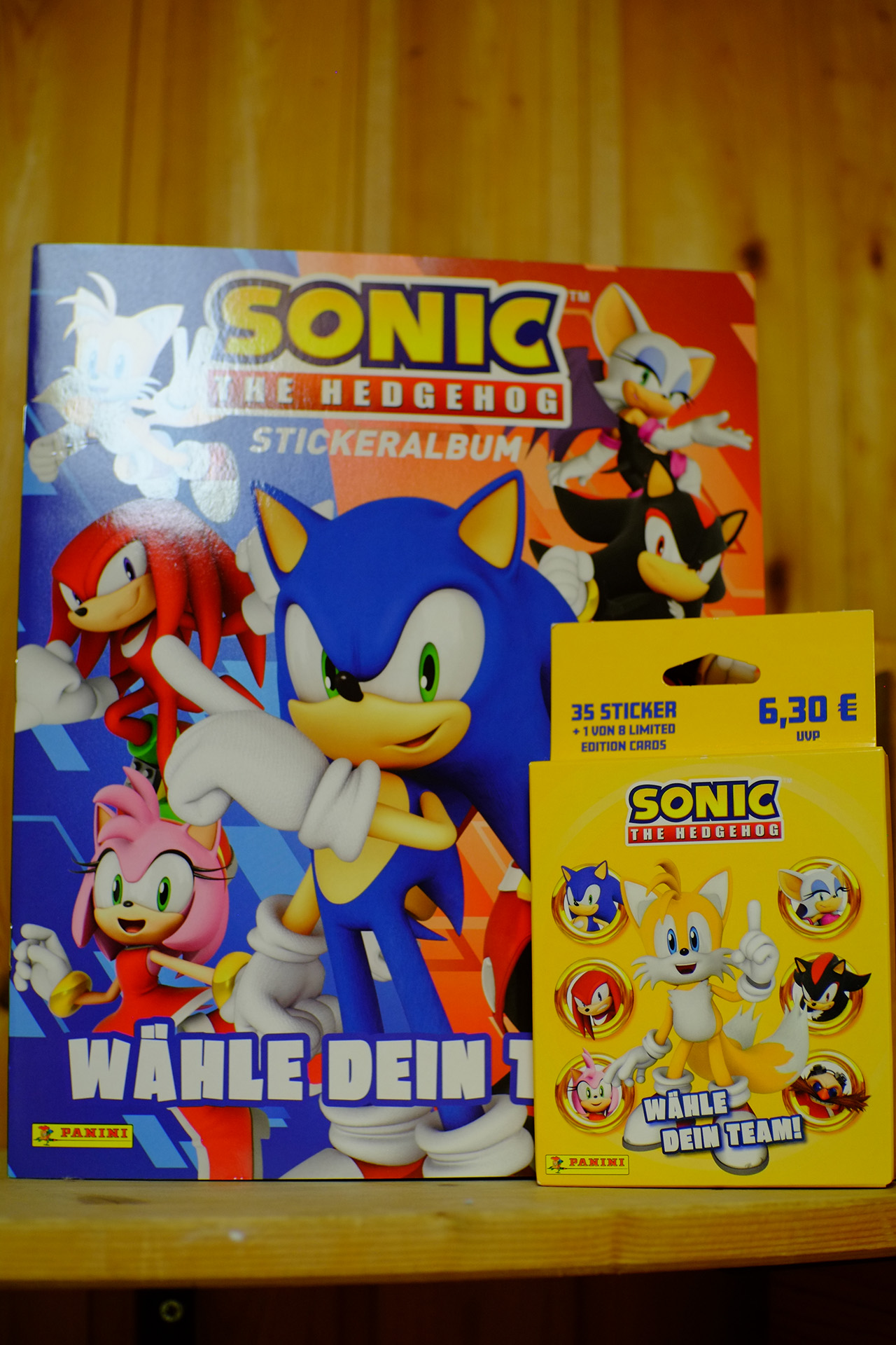 A photo of the Sonic the Hedgehog sticker album and a booster pack for it, which is a small yellow carton box. The sticker album front page features a 3d render of Sonic in the center and 3d renders of Tails, Knuckles, Amy behind Sonic and to the left, and 3d renders of Rouge, Shadow, and Eggman behind Sonic and to the right. The booster pack front art features a 3d render of Tails printed in the center and 3d renders of Sonic, Knuckles, Amy, Rouge, Shadow, and Eggman  are put into golden ring frames to the left and the right of Tails.