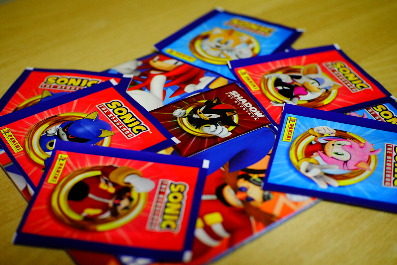 A photo of Sonic the Hedgehog sticker packs organized into a chaotic pile on a table on top of the sticker album, with a foil collectible card featuring Shadow the Hedgehog placed in the center of the pile.