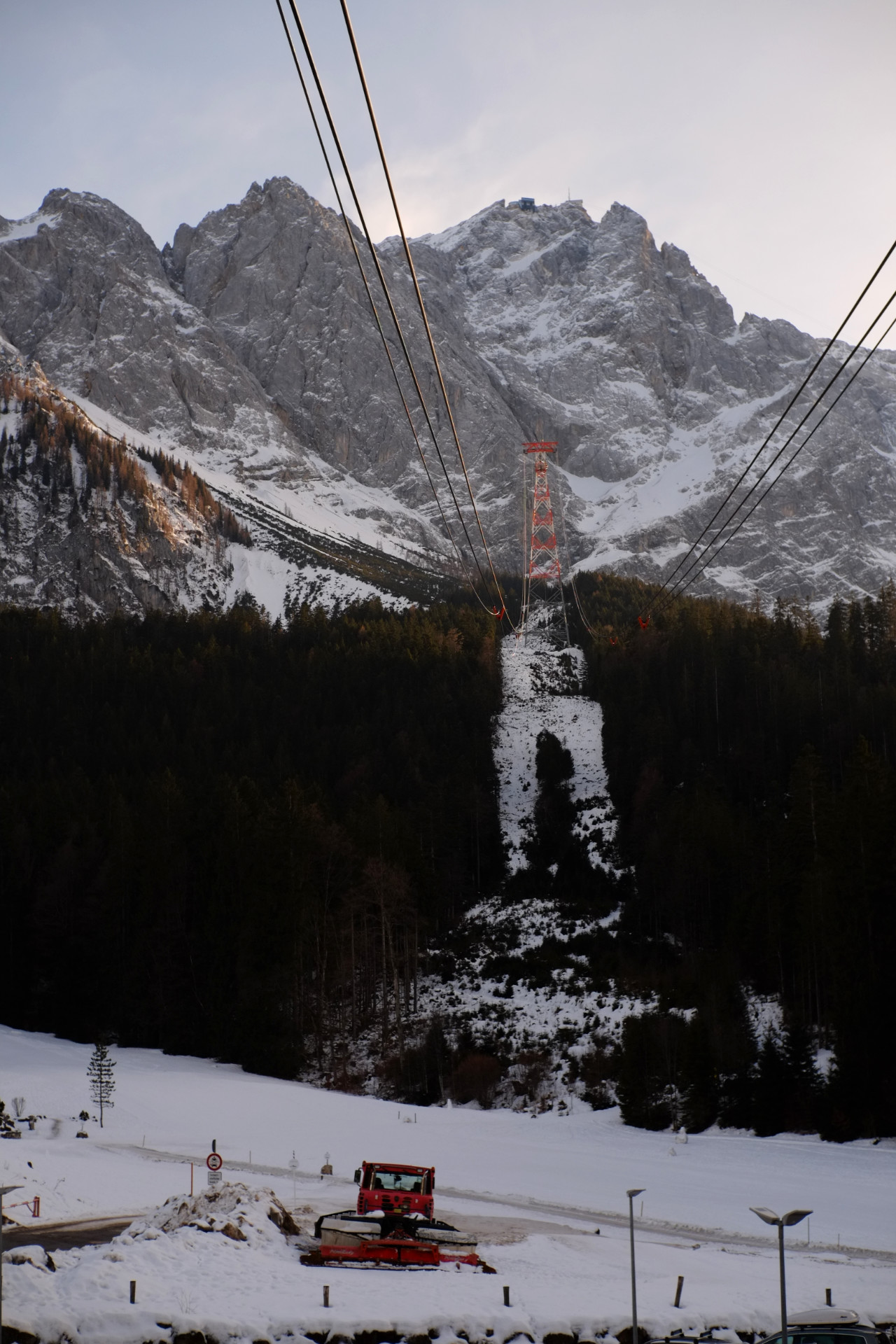 A photo of of cable car cables going up the mountain to the peak station. All the way down is a snowy road with an orange snow plowing car parked nearby.