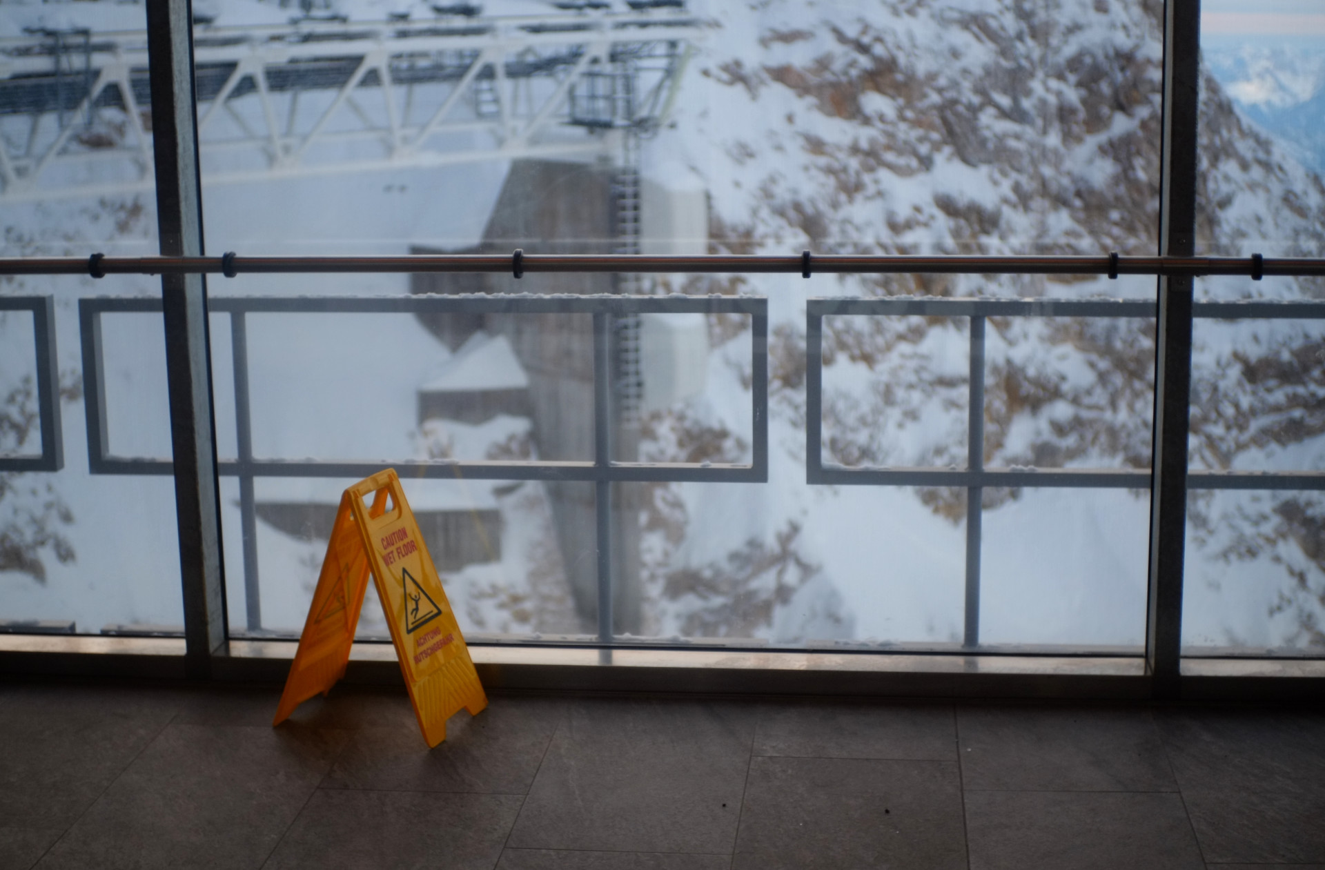 A photo of the cable car peak station hallway with glass walls and a yellow plastic wet floor sign placed on the floor.