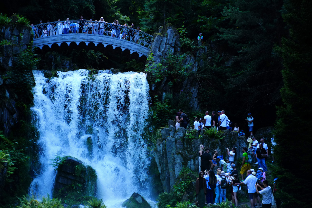 A photo of a waterfall and a metal bridge above it with several tourist on it looking down, and also several tourist climbing a rocky cliff to the right of the waterfall.