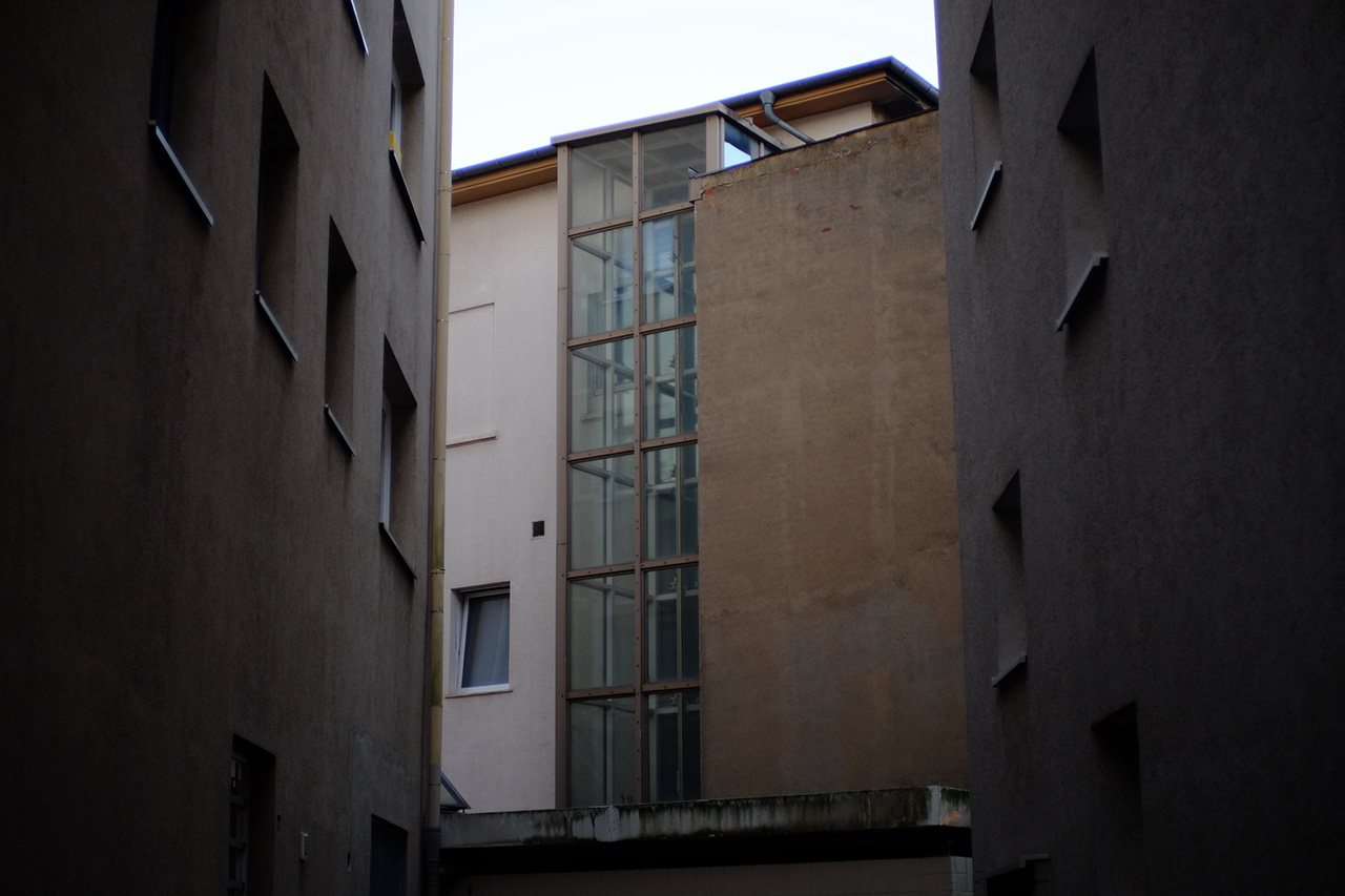 A photo of a glass elevator shaft of a white concrete building with gray concrete walls of two other buildings to the left and to the right.