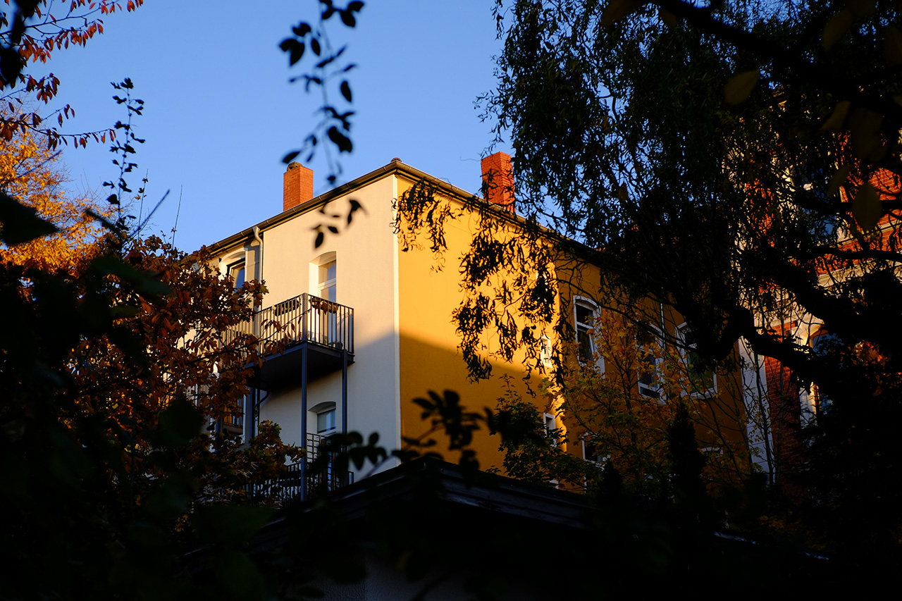 A photo of a low-rise apartment building corner with one wall painted white and another painted yellow. The walls are reflecting sunset light. The view is obstructed by tree branches on the left and on the right.