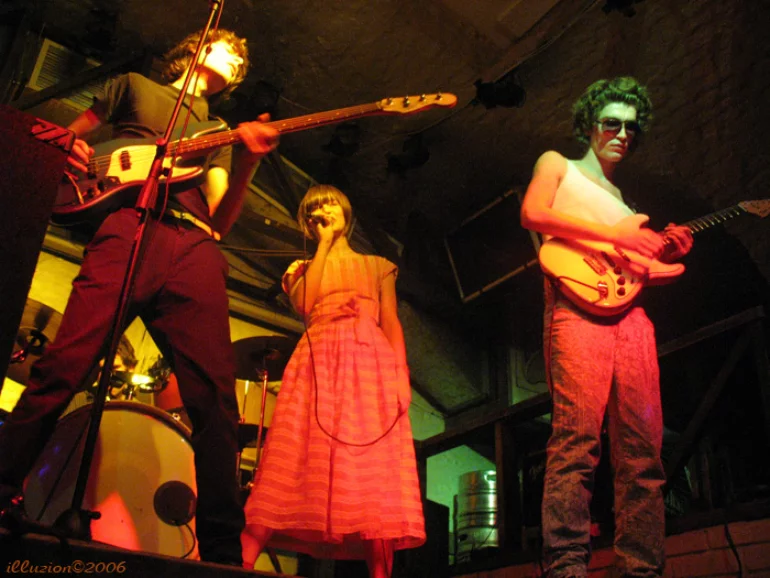 A photo of four people on stage: a vocalist in a long dress in the center, a bass guitarist in a black t-shirt and black trousers, a lead guitarist with dark aviator sunglasses, and a barely visible drummer behind the drums.