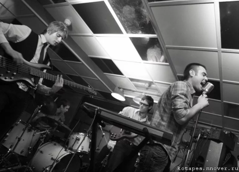 A black-white photo from a stage performance featuring a singer behind a synthesizer, a bass player, a lead guitarist, and a drummer in the background.