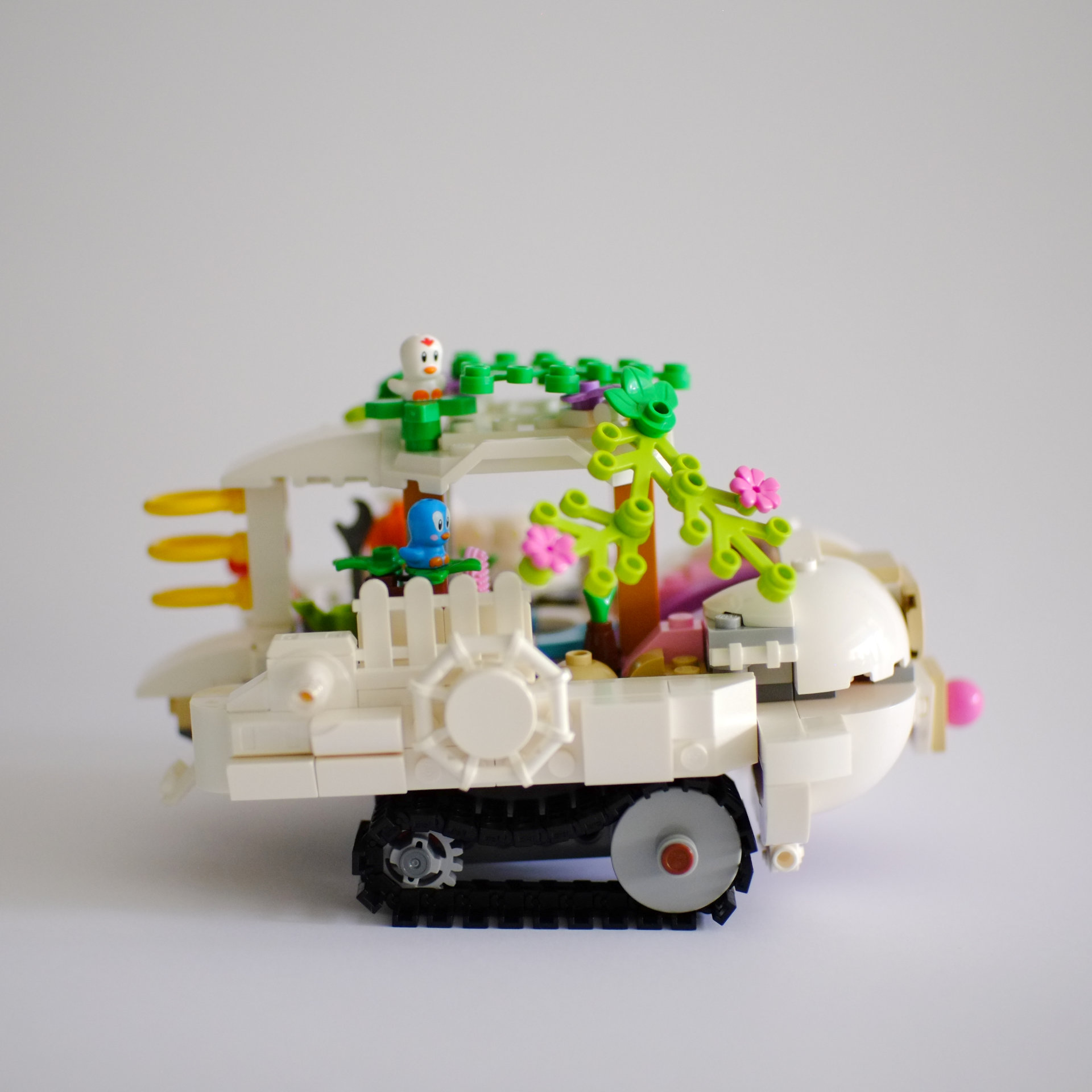 A photo of the right side of a lego build that is a white tourist boat on tank tracks. The roof of the boat is covered with foliage with birds sitting on top.