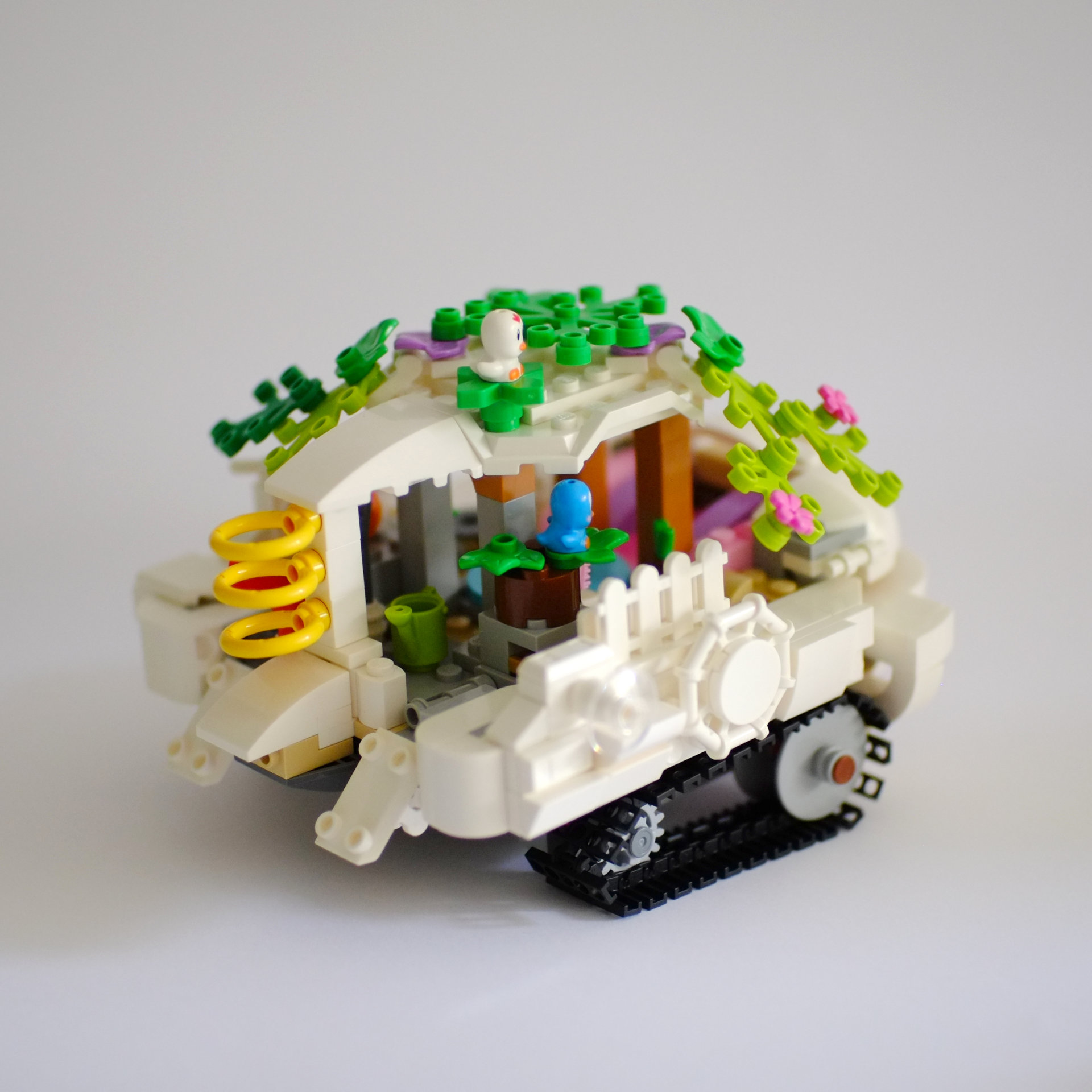A photo of the right side and the back of a lego build that is a white tourist boat on tank tracks. The roof of the boat is covered with foliage with birds sitting on top.