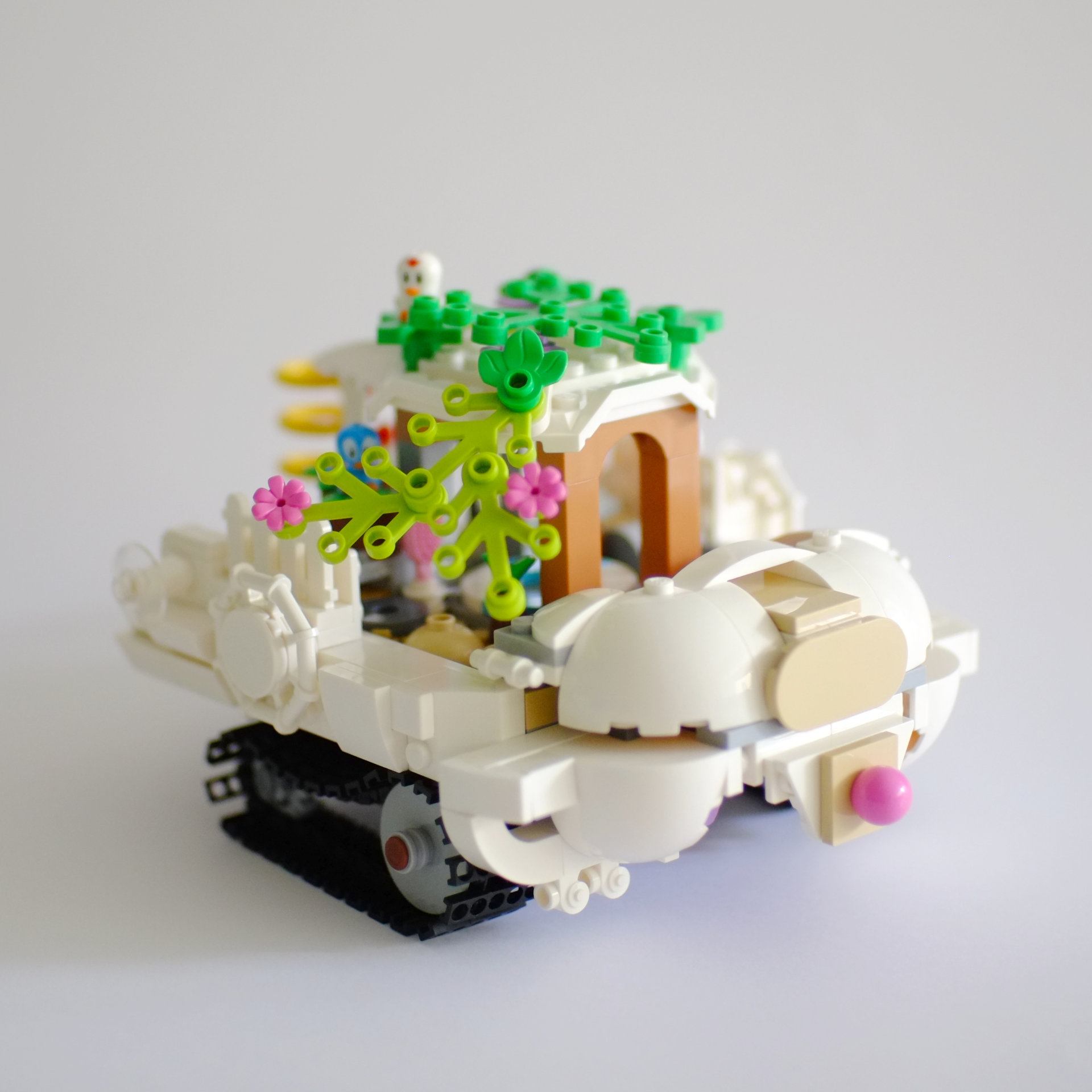 A photo of the right side and the front of a lego build that is a white tourist boat on tank tracks. The roof of the boat is covered with foliage with birds sitting on top.