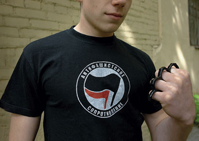 A close-up photo of a person in a black shirt with an Antifascist Movement logo (a white circle with a black and a red flags) showing brass knuckles in his hand.