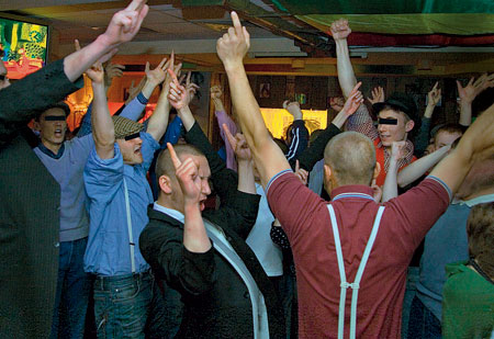 A photo of a group of people, some of them with shaved heads and in suspenders, joyfully raising hands in a bar-looking space.