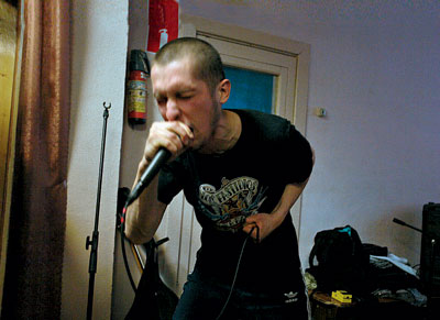 A photo of a man in a black t-shirt, black pants, with his head shaved, holding a microphone in one hand, a microphone cable in another, and singing with his eyes closed.